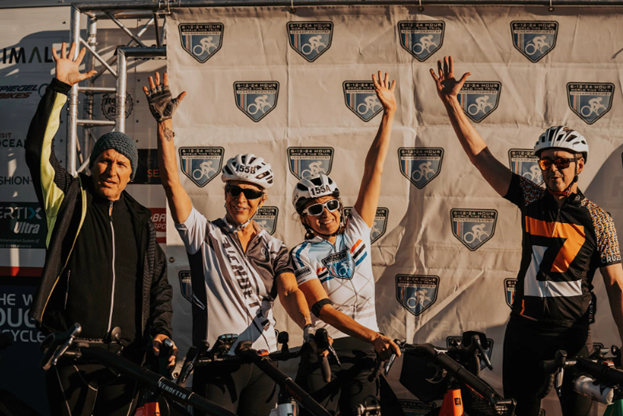 4 bicyclists standing by their bikes with arms in the air.