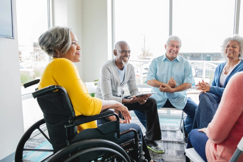 A group of older adults sits in a circle in a bright, welcoming room, engaging in a support group discussion.