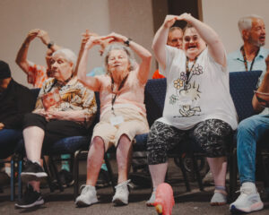 Group of Crescendo Choir members from the West Valley Life Enrichment Program seated and enthusiastically participating in a musical performance, with some forming arm motions above their heads and smiling.