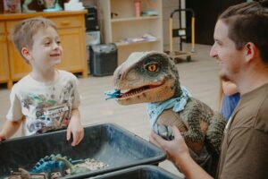 Smiling young boy interacts with a playful dinosaur puppet held by an adult during a themed educational activity at Wirtzie’s Preschool, surrounded by dinosaur toys in sensory bins.