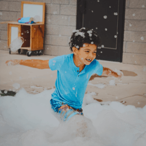 Smiling young child in a blue shirt joyfully running through a pile of foam bubbles outdoors during a playful activity at Wirtzie’s Child Care.