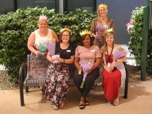 From left to right: Courtney Allen (VP of Program Operations), Lindsey Nichols (Director, West Valley Life Enrichment), Lakisha Shavers (Director, Mary’s Place Life Enrichment), Lisa Minette (Senior Director of Enrichment), and Dawna Gallant (Director, Lucy Anne’s Place Life Enrichment) sit on a bench outdoors wearing paper crowns and holding bouquets, celebrating Benevilla’s 7th consecutive CARF Three-Year Accreditation.