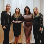 Joanne Byers, Oana Constantinescu, Catherine Lamirato, and Sara Villanueva stand together smiling as Oana holds the Business Leadership Award on behalf of FirstBank at the Benevilla Gratitude Celebration Lunch.