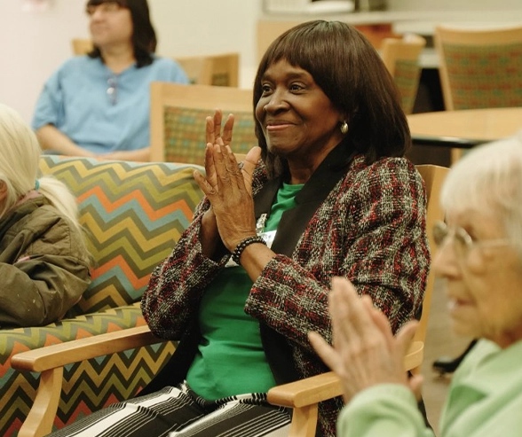 Older adult woman seated at Mary’s Place Life Enrichment adult day program, smiling and clapping during a group activity, with other participants in the background engaged in the session.