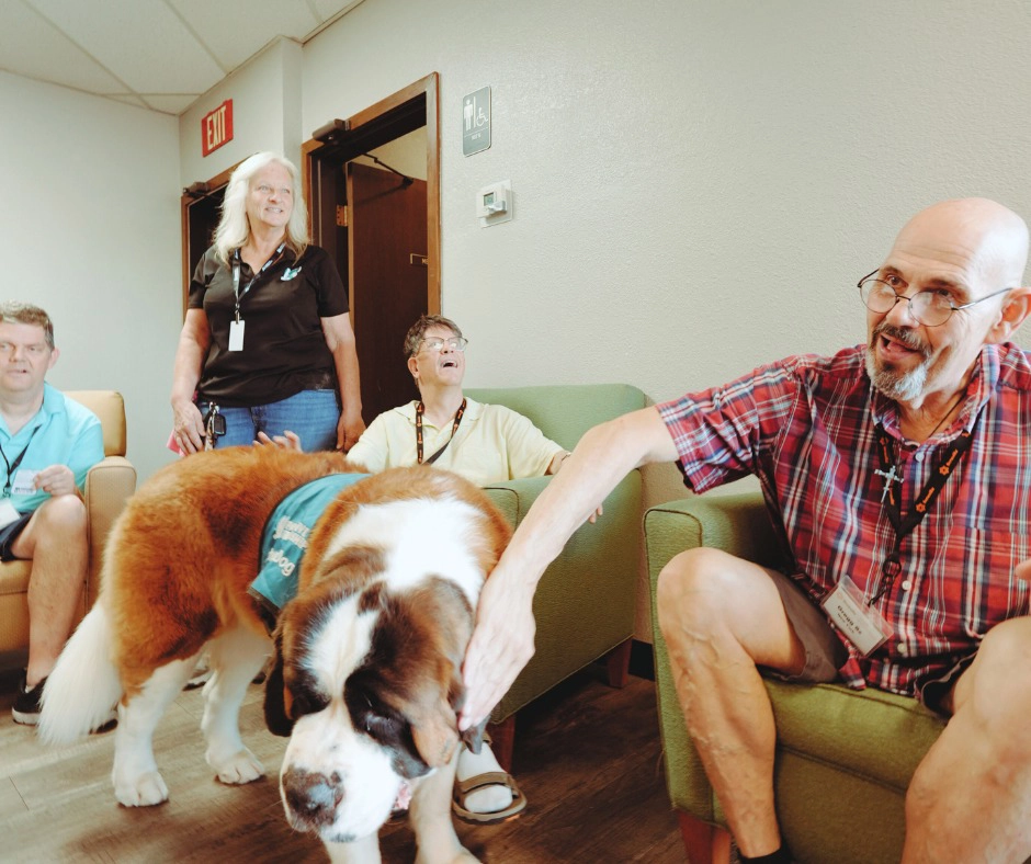 Adult day program participants sit and smile in a lounge area as a large St. Bernard therapy dog is gently petted, with a staff member standing nearby during an animal assisted activity at Benevilla’s West Valley Life Enrichment Program.